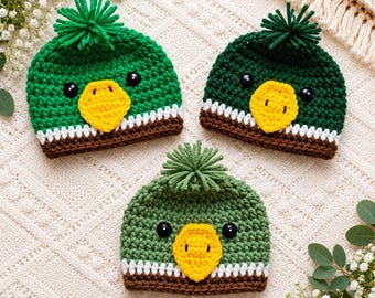 May include: Three crocheted beanies designed to resemble mallard ducks. Each hat features a green crown with a yarn pom-pom on top, black button eyes, a yellow beak, and a white and brown striped band along the bottom edge of the headwear.