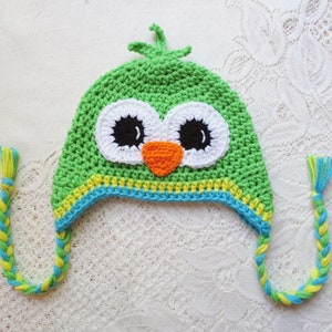 Puede incluir: Un gorro de b&uacute;ho verde tejido a ganchillo con rasgos faciales blancos, negros y naranjas. El gorro tiene una banda a rayas amarillas y azules y lazos trenzados con borlas amarillas y azules. El gorro es para ni&ntilde;os.