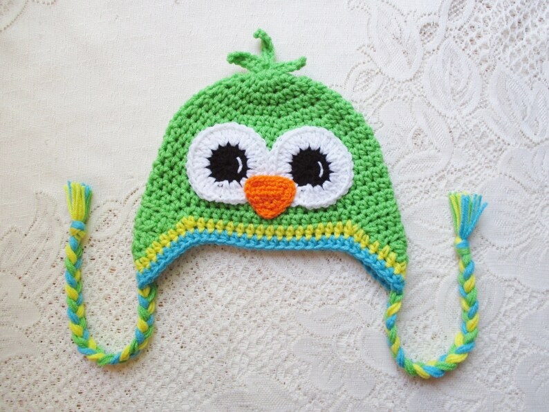 Puede incluir: Un gorro de b&uacute;ho verde tejido a ganchillo con rasgos faciales blancos, negros y naranjas. El gorro tiene una banda a rayas amarillas y azules y lazos trenzados con borlas amarillas y azules. El gorro es para ni&ntilde;os.