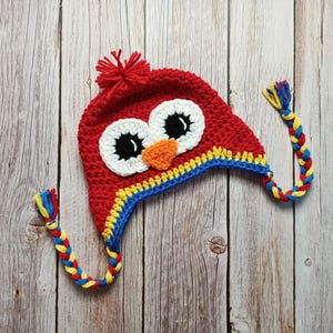 Puede incluir: Un gorro rojo de ganchillo con un dise&ntilde;o de b&uacute;ho. El gorro presenta grandes ojos blancos y negros, un pico naranja y un ribete amarillo y azul. Borlas trenzadas en amarillo, azul y rojo cuelgan de los lados. Un pomp&oacute;n rojo est&aacute; en la parte superior.