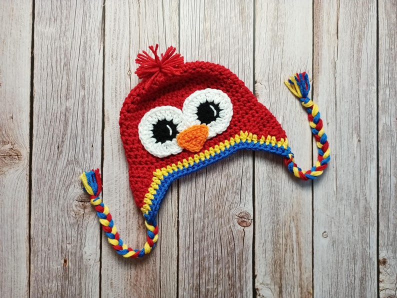 Puede incluir: Un gorro rojo de ganchillo con un dise&ntilde;o de b&uacute;ho. El gorro presenta grandes ojos blancos y negros, un pico naranja y un ribete amarillo y azul. Borlas trenzadas en amarillo, azul y rojo cuelgan de los lados. Un pomp&oacute;n rojo est&aacute; en la parte superior.