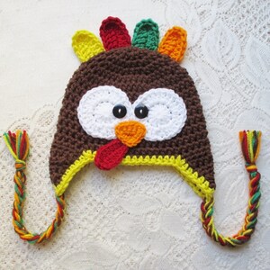 May include: A brown crocheted turkey hat with a yellow brim, white eyes, an orange beak, and a red wattle. The hat has long, colourful braided ties.