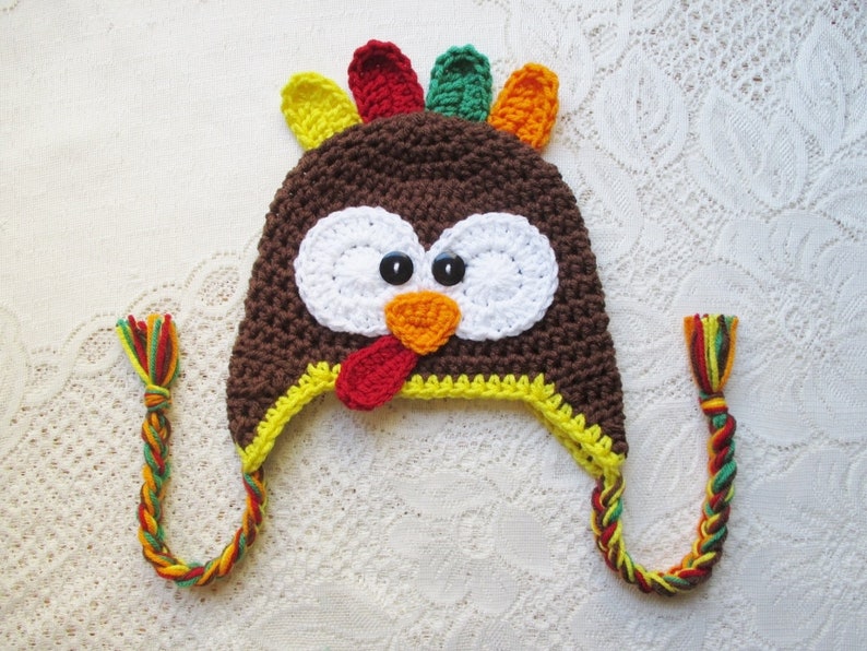 May include: A brown crocheted turkey hat with a yellow brim, white eyes, an orange beak, and a red wattle. The hat has long, colourful braided ties.