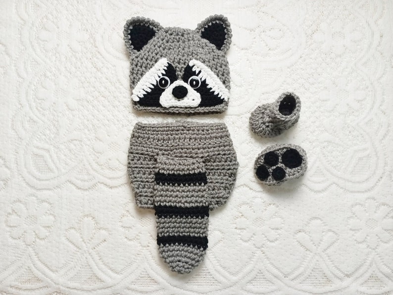 K&ouml;nnte beinhalten: Ein graues und schwarzes H&auml;kel-Waschb&auml;r-Kost&uuml;m-Set f&uuml;r ein Baby, einschlie&szlig;lich Hut, Windel&uuml;berhose und Babyschuhe. Der Hut hat ein Waschb&auml;rgesicht mit wei&szlig;en und schwarzen Details. Die Windel&uuml;berhose hat einen gestreiften Schwanz, und die Babyschuhe haben Pfoten.