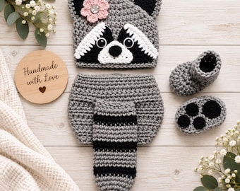 May include: A crochet raccoon set including a hat with a pink flower, a diaper cover with an attached striped tail, and two booties. The hat features black and white facial details and round eyes. One bootie has black paw pad accents.