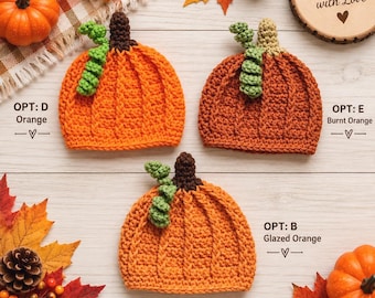 Pode incluir: Três gorros de crochê em forma de abóbora com caules verdes encaracolados e hastes castanhas ou bege. Os gorros estão rotulados como Opt: D Orange, Opt: E Burnt Orange e Opt: B Glazed Orange. O texto Handmade with Love está num círculo de madeira.