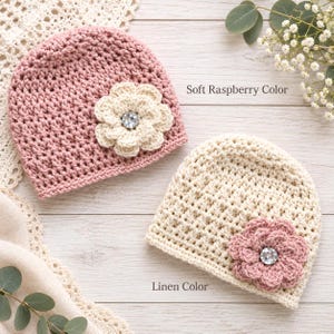 May include: Two crocheted beanies, one in soft raspberry with a cream-colored flower and one in linen with a raspberry-colored flower. Each flower features a central clear rhinestone. The text Soft Raspberry Color and Linen Color is visible.