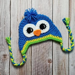 Puede incluir: Un gorro de b&uacute;ho azul tejido a ganchillo con ojos blancos, pupilas negras y un pico naranja. El gorro tiene un pomp&oacute;n azul en la parte superior y orejeras trenzadas en amarillo y verde. El gorro est&aacute; hecho a mano y est&aacute; sobre una superficie de madera.