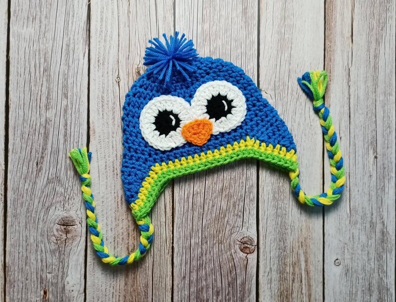 Puede incluir: Un gorro de b&uacute;ho azul tejido a ganchillo con ojos blancos, pupilas negras y un pico naranja. El gorro tiene un pomp&oacute;n azul en la parte superior y orejeras trenzadas en amarillo y verde. El gorro est&aacute; hecho a mano y est&aacute; sobre una superficie de madera.