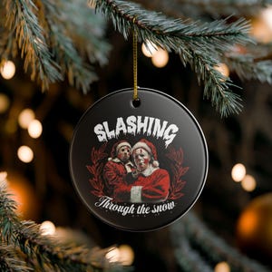 Könnte beinhalten: Schwarzer runder Weihnachtsschmuck mit dem Text "SLASHING" über einer Illustration von zwei Figuren in Weihnachtsmann-Outfits. Der Text "Through the snow" steht darunter. Der Schmuck hängt an einer goldenen Schnur.