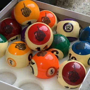 Könnte beinhalten: Ein Satz Billardkugeln in einer weißen Box. Die Kugeln sind in verschiedenen Farben, darunter Orange, Rot, Grün und Blau, mit weißen und schwarzen Akzenten. Jede Kugel hat eine Nummer und eine Handgesten-Grafik.