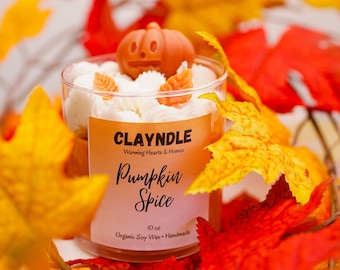 May include: A clear glass candle with white wax, topped with a pumpkin and leaf-shaped decorations. The label reads "Pumpkin Spice" and "10 oz Organic Soy Wax Handmade". Surrounded by orange, red, and yellow fall leaves.