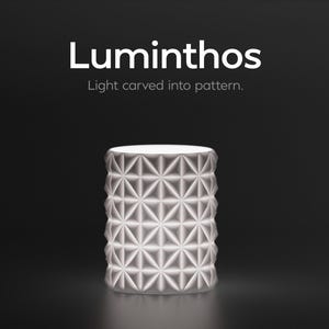 Könnte beinhalten: Eine zylindrische, weiße Lampe mit einem geometrischen Muster aus Dreiecken und Rauten. Die Lampe wird von innen beleuchtet, wodurch ein sanftes Leuchten entsteht. Der Text "Luminthos" und "Light carved into pattern" wird über der Lampe vor schwarzem Hintergrund angezeigt.
