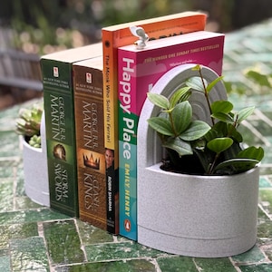 Peut inclure: Deux serre-livres gris clair en forme d'arche à texture pierre contenant quatre livres : A Storm of Swords, A Clash of Kings, The Monk Who Sold His Ferrari et Happy Place. Une plante verte pousse dans le serre-livre droit, surmonté d'un petit oiseau blanc.