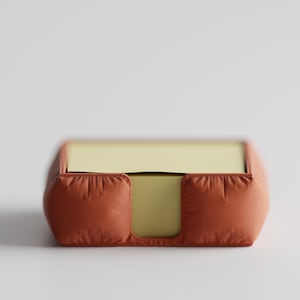 Könnte beinhalten: Ein quadratischer, orangebrauner Notizblockhalter mit weichem, gepolstertem Aussehen. Der Halter enthält einen Stapel gelber Haftnotizen. Das Design ist schlicht und modern, passend für ein Büro oder einen Arbeitsplatz zu Hause.