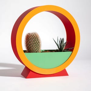 Puede incluir: Una maceta moderna y colorida con un diseño circular abierto. La maceta tiene una base roja, un recipiente verde y un anillo naranja y rojo vibrante. Contiene un cactus y una planta cebra, creando un contraste llamativo.