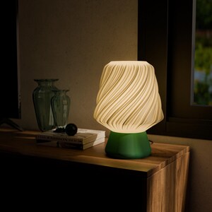May include: A modern table lamp with a cream-colored, swirled shade and a green base. The lamp is turned on, emitting a warm glow. It sits on a wooden surface, next to a window and decorative vases. The lamp's design is contemporary.