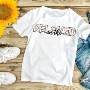 May include: White t-shirt with the words "BELOVED in the" printed across the front. The word "BELOVED" is in a floral pattern, and the words "in the" are in a cursive font. The shirt is paired with white sneakers and blue jeans.