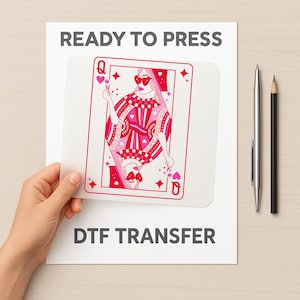 May include: A DTF transfer sheet with a pink Queen of Hearts playing card design. The words "READY TO PRESS" and "DTF TRANSFER" are printed on the white background. A hand holds the transfer sheet. A pen and pencil are on the right.