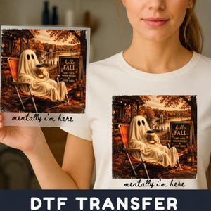 May include: A white t-shirt featuring a graphic of a ghost sitting in a folding chair outdoors, holding a coffee cup. The scene includes a sign that reads "hello FALL COZY DAYS WARM DRINKS & GOOD VIBES" and the text "mentally i'm here" below.