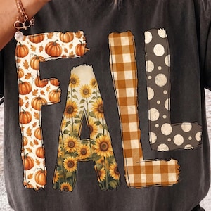 Könnte beinhalten: Ein anthrazitfarbenes T-Shirt mit dem Wort FALL in großen Blockbuchstaben. Das F ist mit orangefarbenen Kürbissen und Blättern gefüllt, das A mit gelben Sonnenblumen, das L mit einem braun-weißen Karomuster und das zweite L mit weißen Punkten.
