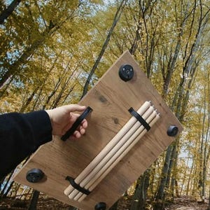 Può includere: Un tavolo in legno con quattro piedini in gomma neri e una maniglia nera. Il tavolo ha una finitura marrone chiaro e include quattro tasselli di legno fissati con cinghie nere. Lo sfondo è una foresta.
