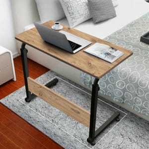 May include: A brown laptop table with black legs and wheels. A laptop, magazine, and coffee cup are on the table. The table is on a grey rug near a bed with white pillows. The table's dimensions are approximately 70cm x 40cm.