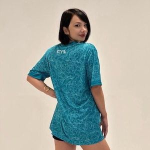 Puede incluir: Camiseta de color turquesa con un estampado floral en espiral. La camiseta de manga corta tiene cuello redondo y la palabra "ATCK" impresa en la espalda. La camiseta se lleva con pantalones cortos negros.