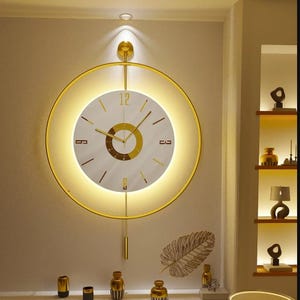 May include: A gold-colored, circular wall clock with a white face and gold hands. The clock has a pendulum and is illuminated with a warm light. The clock is mounted on a light-colored wall, with decorative items below.