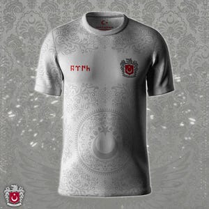 Op de afbeelding: Een wit T-shirt met korte mouwen met een patroon en een rood embleem. Het shirt heeft een wapen met een maan en ster, en tekst in een vreemde taal. Het shirt heeft een ronde hals.