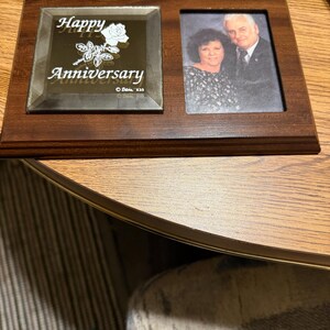 May include: A wooden anniversary picture frame with two sections. One section has a photo of a couple, and the other has the words "Happy Anniversary" with a rose design. The frame is brown and has a hanging loop.