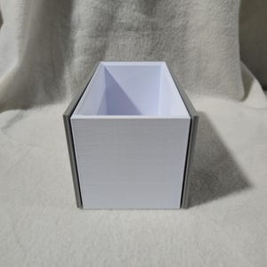 May include: A rectangular container with a white interior and a grey exterior frame. The container is open at the top. It is placed on a white, textured surface. The design is simple and modern.