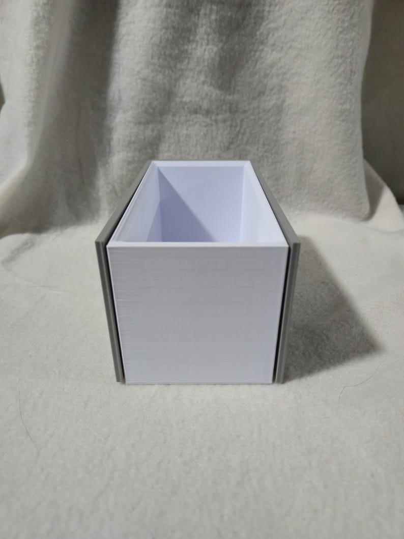 May include: A rectangular container with a white interior and a grey exterior frame. The container is open at the top. It is placed on a white, textured surface. The design is simple and modern.