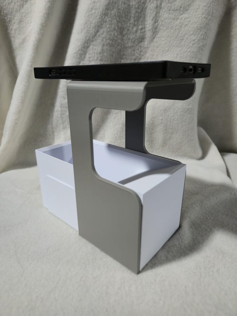 May include: A black rectangular electronic device rests on a grey and white organiser. The organiser has a handle and a white box-shaped base. The background is a soft, off-white fabric.