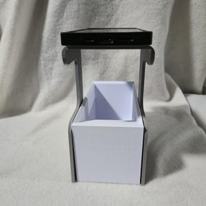 May include: A black smartphone sits on a grey, 3D-printed stand. Below is a rectangular white container, possibly for storage. The stand's design is minimalist and modern, ideal for a desk or bedside table.