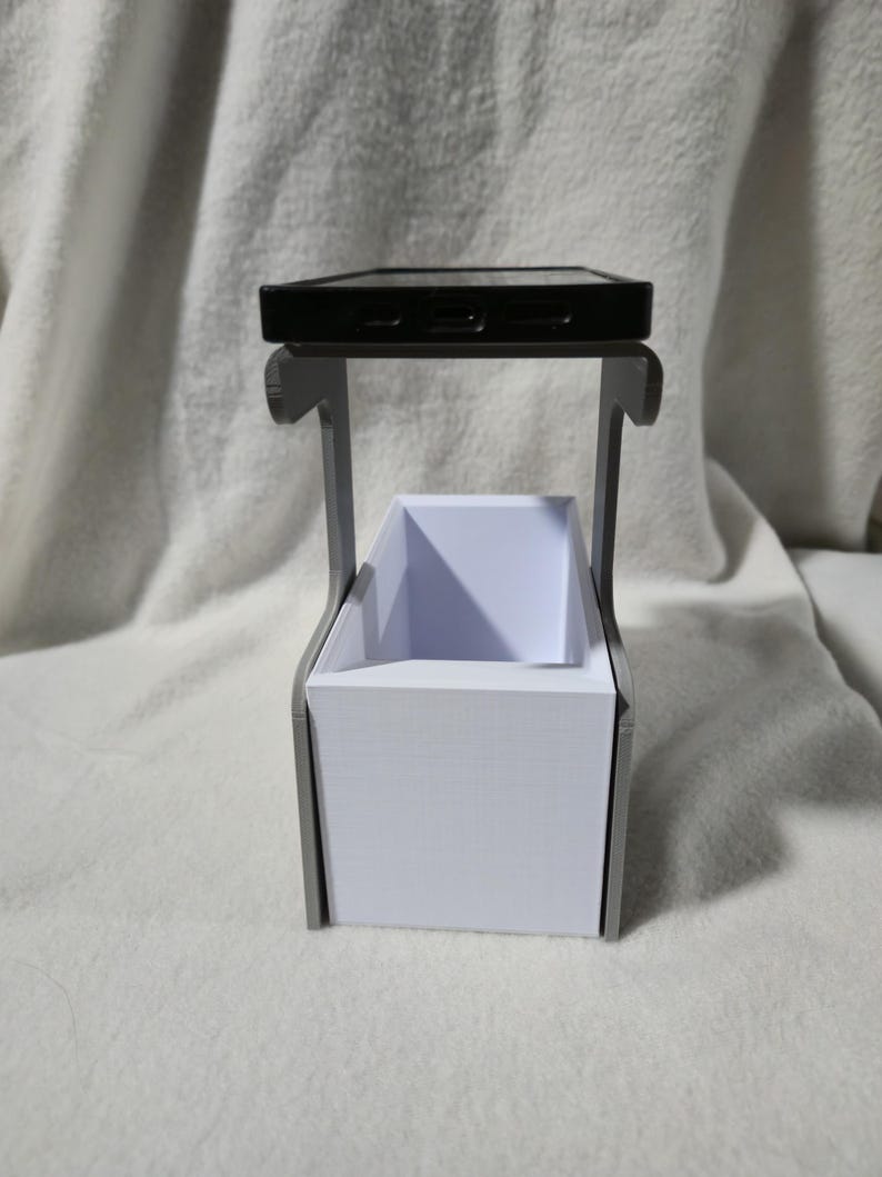 May include: A black smartphone sits on a grey, 3D-printed stand. Below is a rectangular white container, possibly for storage. The stand's design is minimalist and modern, ideal for a desk or bedside table.