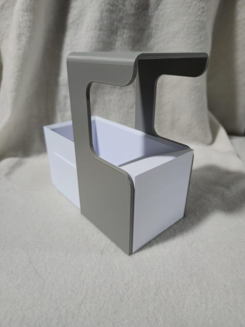May include: A grey and white organiser with a handle. The organiser has a rectangular white box with a grey handle attached to the side. The handle curves over the top of the box. The organiser is on a white surface.