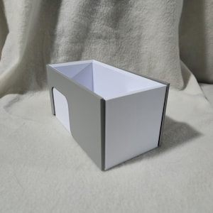 May include: A rectangular storage box with a white interior and grey exterior trim. The box has a cutout on one side and is sitting on a white, textured surface. The box is empty.
