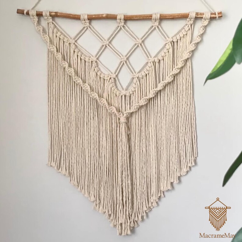 May include: Cream-coloured macrame wall hanging with a geometric design. The wall decor features a wooden dowel at the top, diamond-shaped knots, and a fringed bottom. The piece is handmade and adds a bohemian touch to any room.