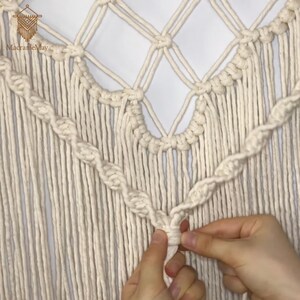 May include: Cream macrame wall hanging with a geometric design. The piece features a diamond pattern at the top and a twisted rope detail. The text "MacrameMay" is visible in the upper left corner. A person's hands are shown working on the macrame.
