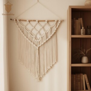 May include: Cream-coloured macrame wall hanging with a geometric diamond pattern and fringe. The wall decor is suspended from a wooden dowel and features the text "MacrameMay". This handcrafted piece adds a bohemian touch to any space.