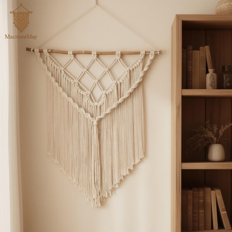 May include: Cream-coloured macrame wall hanging with a geometric diamond pattern and fringe. The wall decor is suspended from a wooden dowel and features the text "MacrameMay". This handcrafted piece adds a bohemian touch to any space.