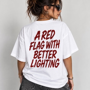 Peut inclure: Un t-shirt blanc à manches courtes avec du texte rouge foncé imprimé au dos. Le texte indique A RED FLAG WITH BETTER LIGHTING dans une police de style pinceau vieilli. Le t-shirt présente un col rond et une coupe ample et décontractée.