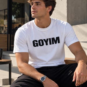May include: A white short-sleeved t-shirt featuring the word GOYIM printed in bold black capital letters across the chest. The garment has a classic crew neckline and a standard fit, constructed from a plain white fabric.