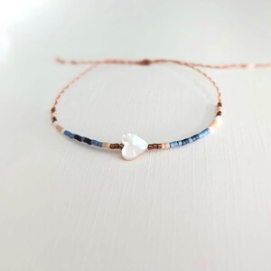 May include: A delicate bracelet featuring a white heart-shaped charm. The bracelet is strung with small, colorful beads in shades of blue, tan, and brown. The cord is a light brown color.
