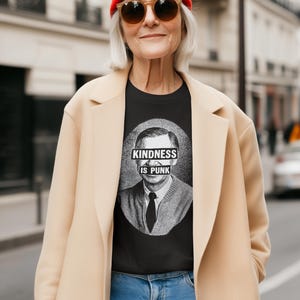 May include: Black t-shirt with a graphic of a man and the text "KINDNESS IS PUNK". The person in the image is wearing a red beanie, sunglasses, a tan coat, and blue jeans.