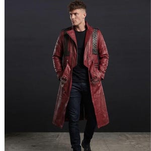 May include: A long, burgundy leather coat with a notched collar, shoulder accents, and buckle details. The coat features a double-breasted design and a knee-length hem. The model is wearing a black shirt and dark blue jeans.