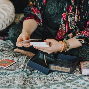 May include: A person holding tarot cards, with a blue box and a deck of cards visible. The person is wearing a floral patterned top and bracelets. The scene is set on a patterned blanket.