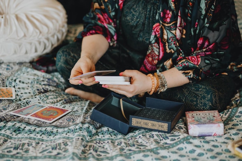 May include: A person holding tarot cards, with a blue box and a deck of cards visible. The person is wearing a floral patterned top and bracelets. The scene is set on a patterned blanket.