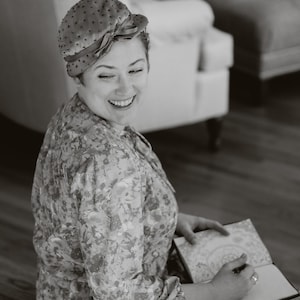 May include: A woman wearing a floral print dress and a dotted hat with a bow, smiling while writing in a notebook. The image is in black and white, with a soft focus, suggesting a vintage or artistic style.